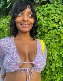 Filmmaker and Poet Jeanetta Rich poses in front of a wall of leaves. She looks off to the left slightly and has a gentle smile. She is wearing a purple blouse with a psychedelic horizontal pattern.